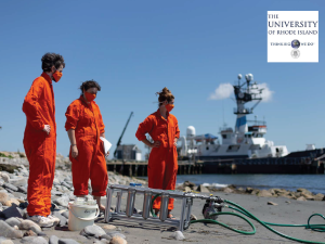 photo of three people in orange jumpsuits and facemasks on a shore with pump equipment and a sea vessel in the background