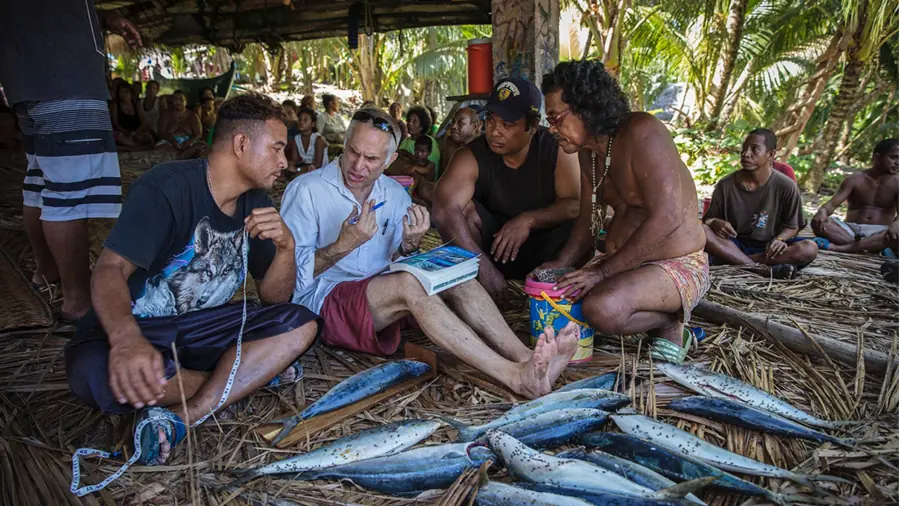 seated people talking under a structure next to fish catch laid out in front of them, many people in the background